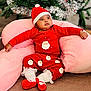 baby, child, christmas, holiday, festive_clothing, red_pajamas, santa_hat, pink_cushion, indoor, decorated_tree, greenery, celebration, cute, infant, smiling, soft_texture, holiday_season, cozy, portrait, relaxed
