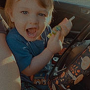Emmitt is registered to the contest to win money with this photo: child, toddler, car_interior, steering_wheel, blue_shirt, plaid_shorts, sunlight, happy, smiling, toy, hand, seat, window, daylight, mushroom_pattern, person, vehicle, casual_clothing, fun, expression