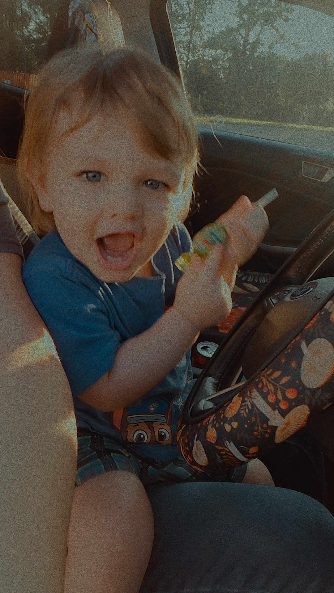 Emmitt is registered to the contest to win money with this photo: child, toddler, car_interior, steering_wheel, blue_shirt, plaid_shorts, sunlight, happy, smiling, toy, hand, seat, window, daylight, mushroom_pattern, person, vehicle, casual_clothing, fun, expression