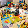 toddler, child, toy, playmat, indoor, colorful, puzzle_mat, floor, plastic_toys, baby, child_playing, furniture, radiator, box, floor_tiling, smiling, cute, seated, blue_clothing, playroom