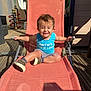 toddler, child, smiling, outdoor, chair, blue_tank_top, striped_shorts, shoes, wooden_deck, sunlight, shadow, toy, playful, happy, summer, relaxing, furniture, person, casual, daytime