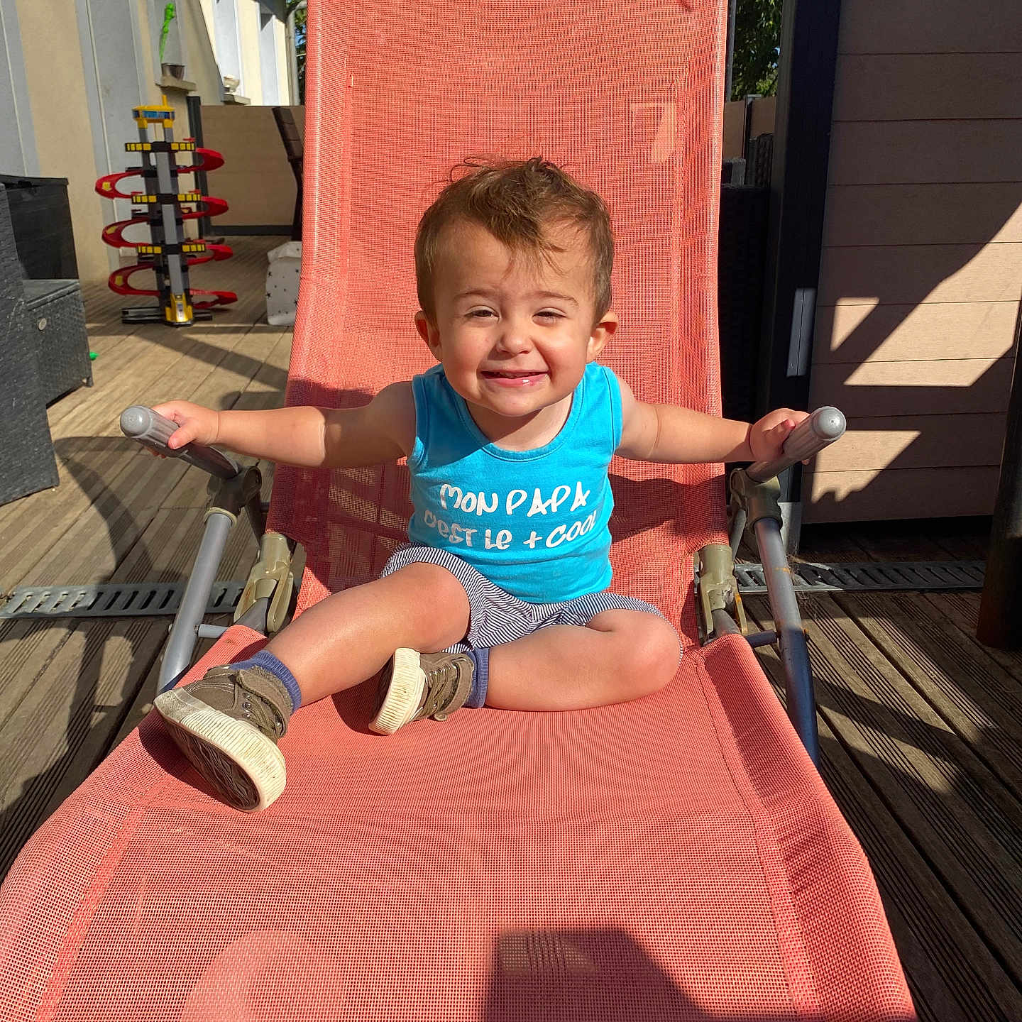 Gabriel a rejoint le concours — aidez-le/la à gagner de superbes lots ! blue_tank_top, casual, chair, child, daytime, furniture, happy, outdoor, person, playful, relaxing, shadow, shoes, smiling, striped_shorts, summer, sunlight, toddler, toy, wooden_deck