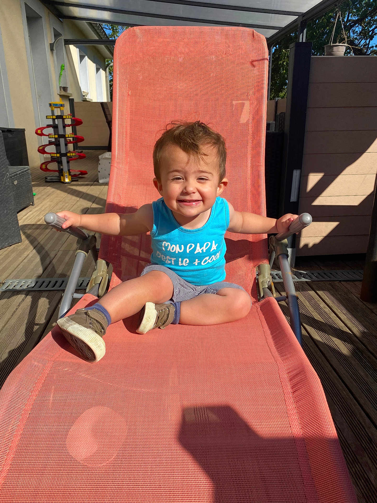 Gabriel a rejoint le concours — aidez-le/la à gagner de superbes lots ! toddler, child, smiling, outdoor, chair, blue_tank_top, striped_shorts, shoes, wooden_deck, sunlight, shadow, toy, playful, happy, summer, relaxing, furniture, person, casual, daytime