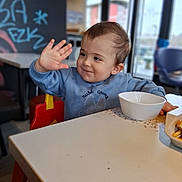 Gabriel participe au concours pour gagner de l'argent avec cette photo : toddler, child, blue_sweatshirt, high_chair, table, bowl, food, indoor, cafe, graffiti, window, daylight, smiling, waving_hand, person, face, hair, seat, casual, happy