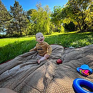 River is registered to the contest to win money with this photo: baby, child, blanket, toy, apple, grass, tree, sunny, outdoor, nature, greenery, smiling, happy, playing, park, blue_sky, daylight, person, fall, casual