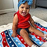 Khyah joined the competition — help win amazing prizes! baby, child, infant, smiling, happy, portrait, indoors, bedroom, headband, bow, red_outfit, blue_accessory, blanket, stars, patriotic, sitting, carpet, dresser, sandals, family_photo