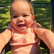 Khyah joined the competition — help win amazing prizes! baby, child, infant, smiling, happy, portrait, outdoor, sunlight, park, lawn, fence, seated, pink_outfit, dress, ruffled_collar, cherry_detail, curly_hair, brown_eyes, close_up, photography