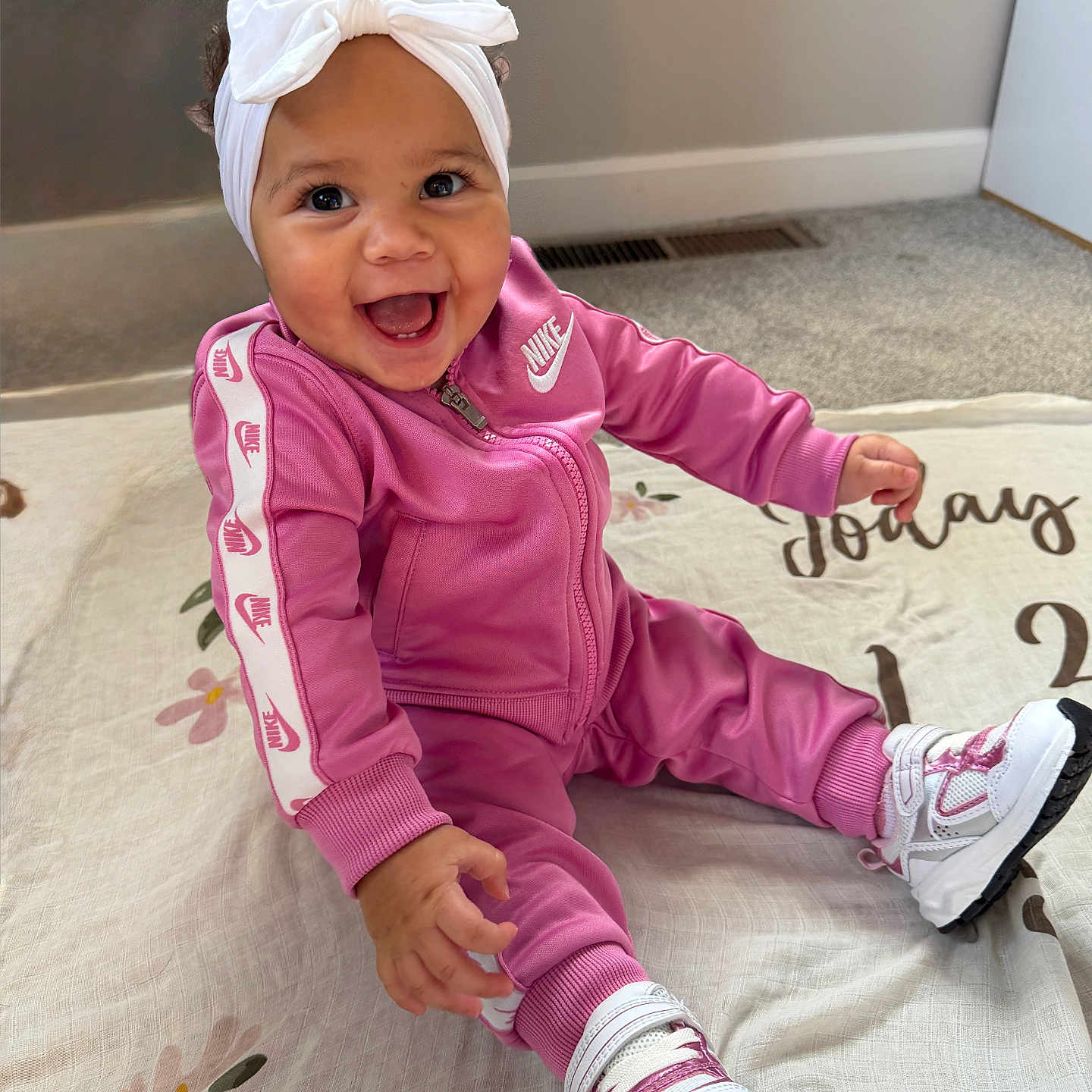 Khyah is registered to the contest to win money with this photo: baby, child, infant, smiling, happy, portrait, sitting, indoor, pink_outfit, tracksuit, headband, bow, sneakers, clothing, blanket, floral_pattern, carpet, home, floor, nike