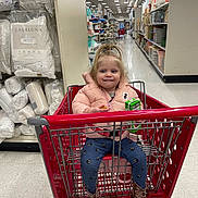 Emilena Joy is registered to the contest to win money with this photo: toddler, shopping_cart, store_aisle, pillows, pink_jacket, blue_pants, brown_boots, juice_box, snack, child, smiling, indoor, floor_tiles, shelves, home_goods, shopping, person, cute, happy, retail