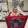 toddler, shopping_cart, store_aisle, pillows, pink_jacket, blue_pants, brown_boots, juice_box, snack, child, smiling, indoor, floor_tiles, shelves, home_goods, shopping, person, cute, happy, retail