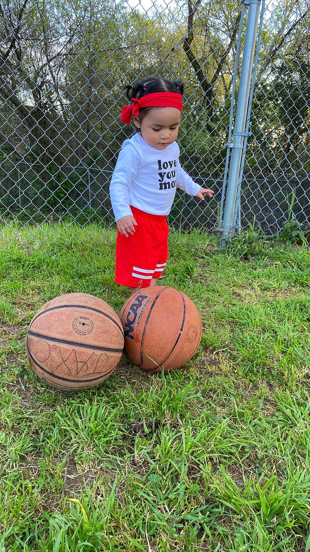 Xitlali is registered to the contest to win money with this photo: ball, ball_game, basketball, chain_link_fencing, child, fence, grass, hat, headwear, mesh, net, people_in_nature, person, plant, player, shorts, sports, sports_equipment, team_sport, toddler