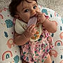 baby, infant, child, cushion, bib, toy, chewing, pink_outfit, animal_pattern, rainbow_pattern, carpet, indoor, cute, playful, curious, soft_toy, lying_down, face, hand, legs