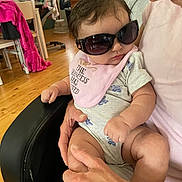 Mary is registered to the contest to win money with this photo: baby, sunglasses, bib, person, armchair, hand, indoor, clothing, infant, cute, child, home, wooden_floor, furniture, pink, holding, casual, relaxed, portrait, expression
