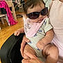 baby, sunglasses, bib, person, armchair, hand, indoor, clothing, infant, cute, child, home, wooden_floor, furniture, pink, holding, casual, relaxed, portrait, expression