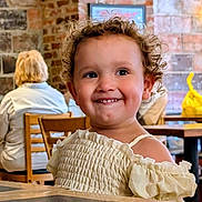Elaine joined the competition — help win amazing prizes! child, toddler, smiling, curly_hair, indoors, cafe, table, brick_wall, wooden_chair, happy, person, clothing, portrait, cute, seated, young_child, dining, light, face, expression