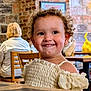 child, toddler, smiling, curly_hair, indoors, cafe, table, brick_wall, wooden_chair, happy, person, clothing, portrait, cute, seated, young_child, dining, light, face, expression