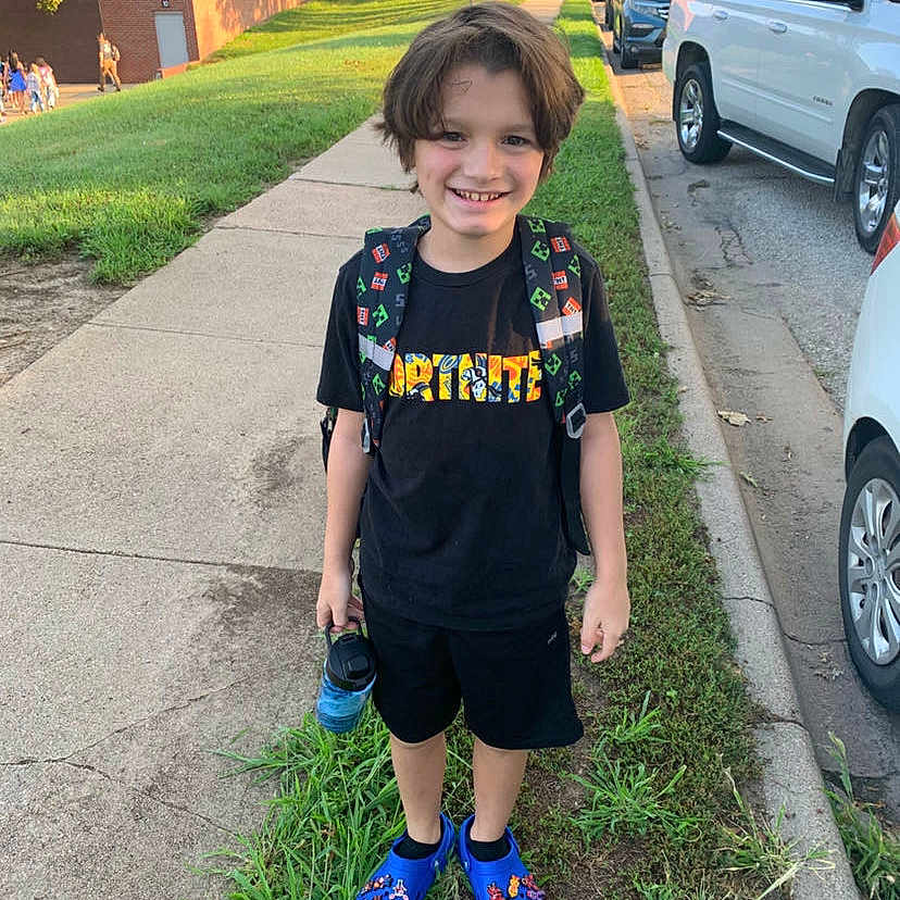 Greycen is registered to the contest to win money with this photo: backpack, blue_shoes, boy, cars, casual_clothing, child, daylight, footwear, grass, happy, outdoor, people_in_background, shorts, sidewalk, smiling, street, trees, tshirt, urban, water_bottle