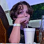child, boy, drink, straw, cup, table, food, pizza, bottle, window, indoor, curly_hair, wristband, black_shirt, face, person, sitting, restaurant, drinkware, casual