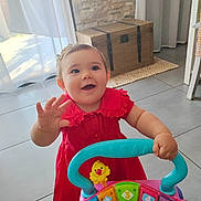 Emma participe au concours pour gagner de l'argent avec cette photo : toddler, child, red_dress, toy, walker, indoor, smiling, happy, play, floor, curtain, wooden_chest, bright_colors, person, young_child, hand_raised, headband, floor_tiles, daylight, home