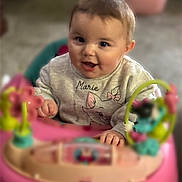 Zada is registered to the contest to win money with this photo: baby, infant, child, smiling, face, eyes, hands, cheeks, hair, sweater, name_on_shirt, toy, walker, pink_toy, play_center, indoors, floor, portrait, cute, playful