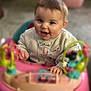 baby, infant, child, smiling, face, eyes, hands, cheeks, hair, sweater, name_on_shirt, toy, walker, pink_toy, play_center, indoors, floor, portrait, cute, playful