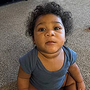 Kaizen is registered to the contest to win money with this photo: baby, child, toddler, sitting, portrait, curly_hair, dark_hair, facial_expression, looking_at_camera, indoors, carpet, home, blue_shirt, short_sleeves, bare_arms, bare_legs, floor, furniture, closeup, photography