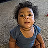 baby, child, toddler, sitting, portrait, curly_hair, dark_hair, facial_expression, looking_at_camera, indoors, carpet, home, blue_shirt, short_sleeves, bare_arms, bare_legs, floor, furniture, closeup, photography
