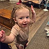 Adalynn is registered to the contest to win money with this photo: toddler, child, indoor, living_room, wooden_floor, couch, braided_hair, smiling, happy, person, baby_gear, playpen, shirt, diaper, floor, wood, home, furniture, human, portrait