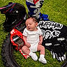 Sutton is registered to the contest to win money with this photo: baby, newborn, sleeping, tire, grass, motocross_helmet, motocross_boots, goggles, gloves, jersey, outdoor, sports_gear, white_clothing, infant, person, resting, protective_gear, black, red, blue