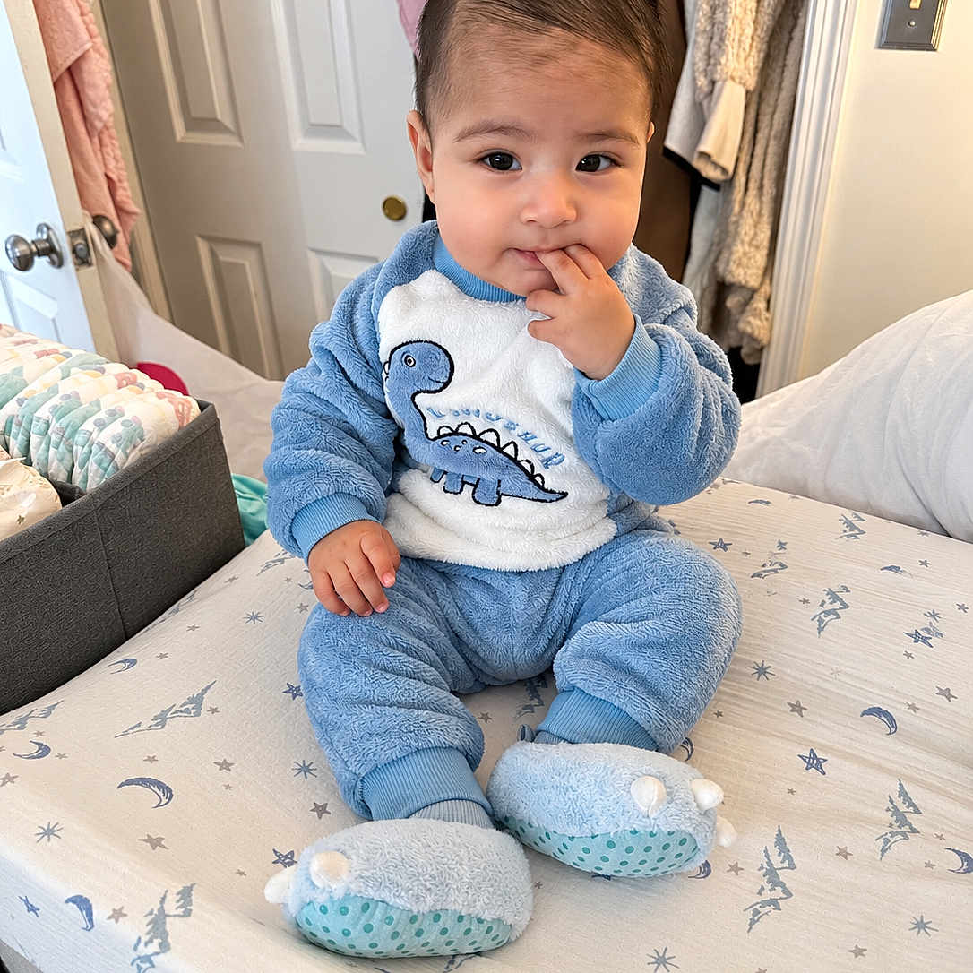 Ethan joined the competition — help win amazing prizes! baby, child, dinosaur_outfit, blue_clothing, sitting, bed, bedroom, portrait, face, smiling, hand_to_mouth, infant, pajamas, slippers, blanket, bedding, closet, indoor, cozy, family_photo