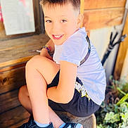 Mathéo participe au concours pour gagner de l'argent avec cette photo : child, boy, smiling, sitting, wooden_stool, shorts, tshirt, sneakers, outdoor, plants, wooden_wall, happy, casual, portrait, young, cute, feet, legs, light, daylight