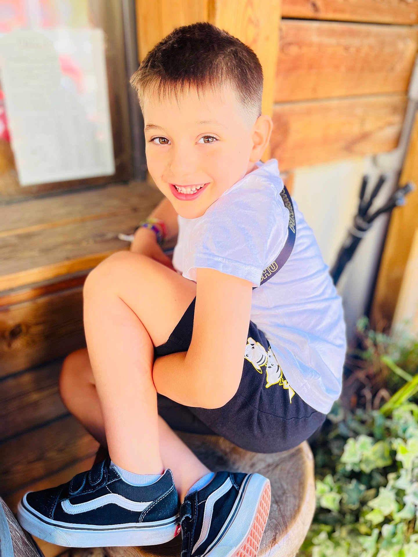 Mathéo participe au concours pour gagner de l'argent avec cette photo : child, boy, smiling, sitting, wooden_stool, shorts, tshirt, sneakers, outdoor, plants, wooden_wall, happy, casual, portrait, young, cute, feet, legs, light, daylight