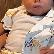 Dajon is registered to the contest to win money with this photo: baby, child, curly_hair, animal_print, clothing, white_shirt, sitting, indoor, cute, portrait, person, hand, skin, young, infant, expression, looking, face, eyes, comfortable