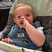 Searra joined the competition — help win amazing prizes! baby, child, high_chair, denim_overalls, messy, chocolate, food, hand, face, blue_eyes, blonde_hair, indoor, curious_expression, smudge, clothing, person, sitting, tray, feeding, cute