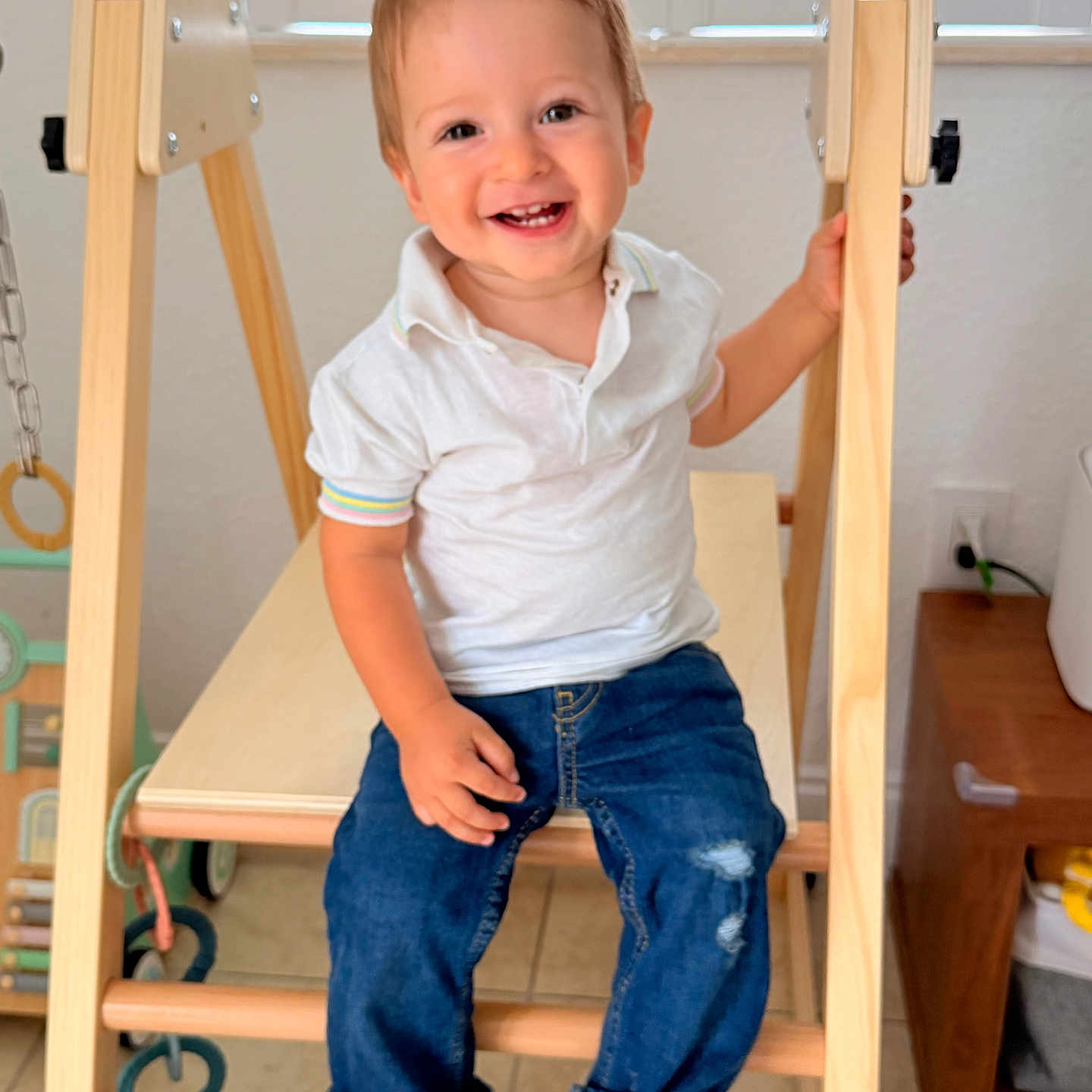 Sebastian is registered to the contest to win money with this photo: barefoot, blue_jeans, child, cute, furniture, happy, home, indoor, jeans, natural_light, person, play, polo_shirt, sitting, smile, toddler, white_shirt, window, wooden_play_structure, young_child