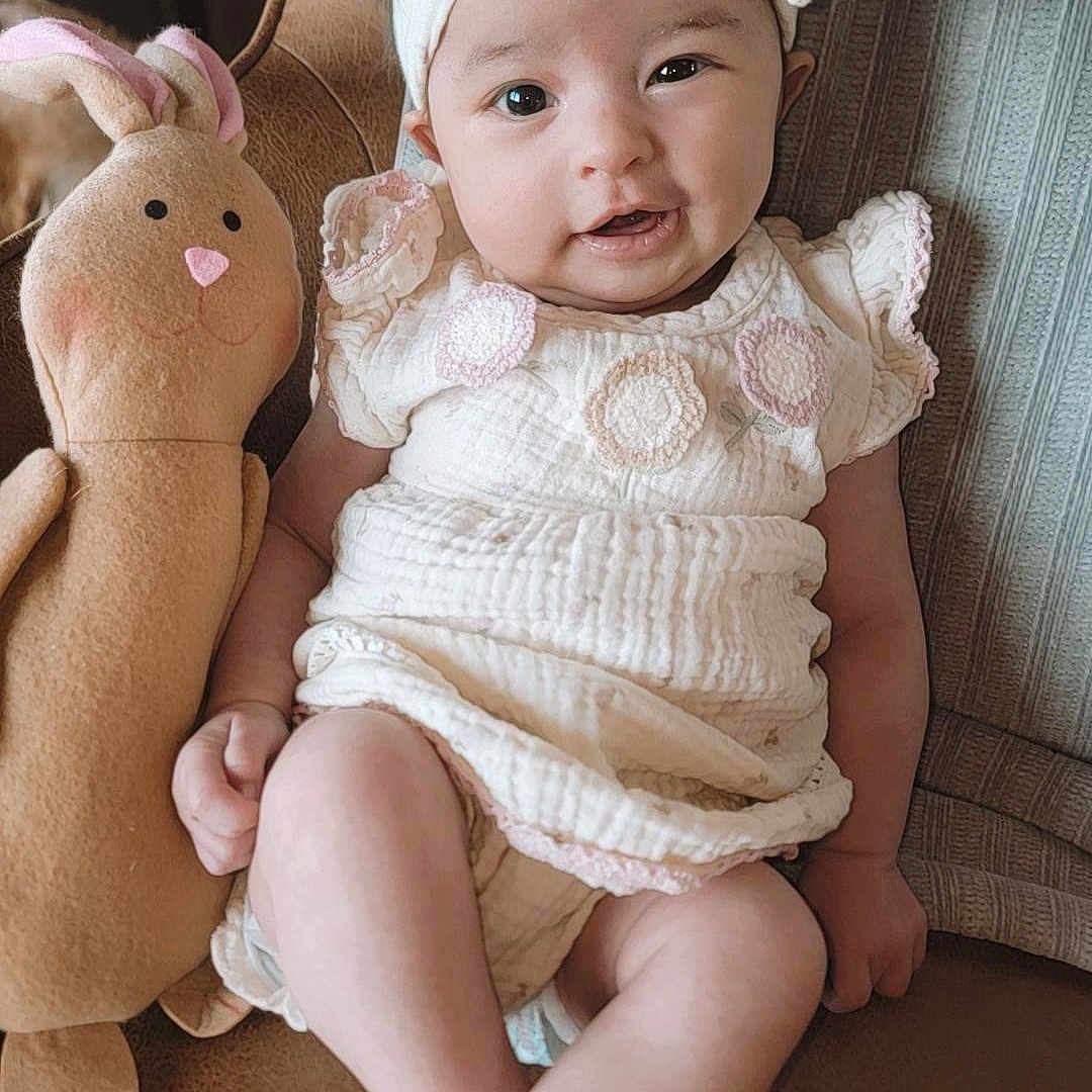 Sloane joined the competition — help win amazing prizes! baby, child, plush_toy, bunny, headband, smile, sitting, dress, indoor, childhood, cute, toy, young_child, person, furniture, cozy, portrait, happy, infant, cuddly