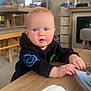 Theo is registered to the contest to win money with this photo: baby, child, indoor, table, book, blue_eyes, curious, black_hoodie, hands, wooden_table, dining_area, chair, curtain, soft_lighting, furniture, face, person, expression, home, cozy