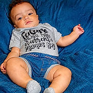John is registered to the contest to win money with this photo: baby, boy, infant, child, tshirt, denim_shorts, blue_blanket, socks, casual_clothing, portrait, indoors, cute, relaxed, skin, face, legs, hand, laying_down, expression, young_child