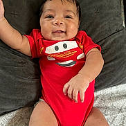 Jasai joined the competition — help win amazing prizes! baby, infant, red_onesie, smiling, happy, face, arm, cushion, blanket, lying_down, cute, child, indoors, skin, person, head, clothing, expression, young, portrait