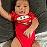 baby, infant, red_onesie, smiling, happy, face, arm, cushion, blanket, lying_down, cute, child, indoors, skin, person, head, clothing, expression, young, portrait
