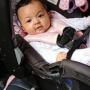 Maura is registered to the contest to win money with this photo: baby, infant, car_seat, straps, safety, plush_toy, curly_hair, clothing, seat, child, car_interior, cute, face, person, indoors, smiling, pink, black, comfort, young