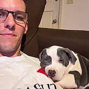 Daisy is registered to the contest to win money with this photo: man, glasses, dog, puppy, couch, white, black, indoor, door, relaxing, cute, pet, animal, face, human, casual, resting, comfort, smile, home