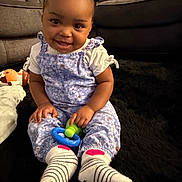 Brooklyne is registered to the contest to win money with this photo: baby, black_rug, child, couch, cozy, cute, floral_jumpsuit, happy, home, indoor, person, pigtails, playful, plush_toy, portrait, seated, smiling, striped_socks, toddler, toy