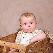 Frankie is registered to the contest to win money with this photo: baby, child, basket, blue_eyes, curly_hair, cute, indoor, neutral_background, wooden_floor, seated, portrait, rosy_cheeks, clothing, patterned_shirt, wicker, person, infant, young_child, looking_away, soft_lighting