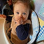 baby, infant, child, high_chair, thumb_sucking, blue_eyes, fuzzy_vest, ponytail, bib, tray, patterned_fabric, indoor, jacket, backpack, dining_area, face, portrait, close_up, curious, seated