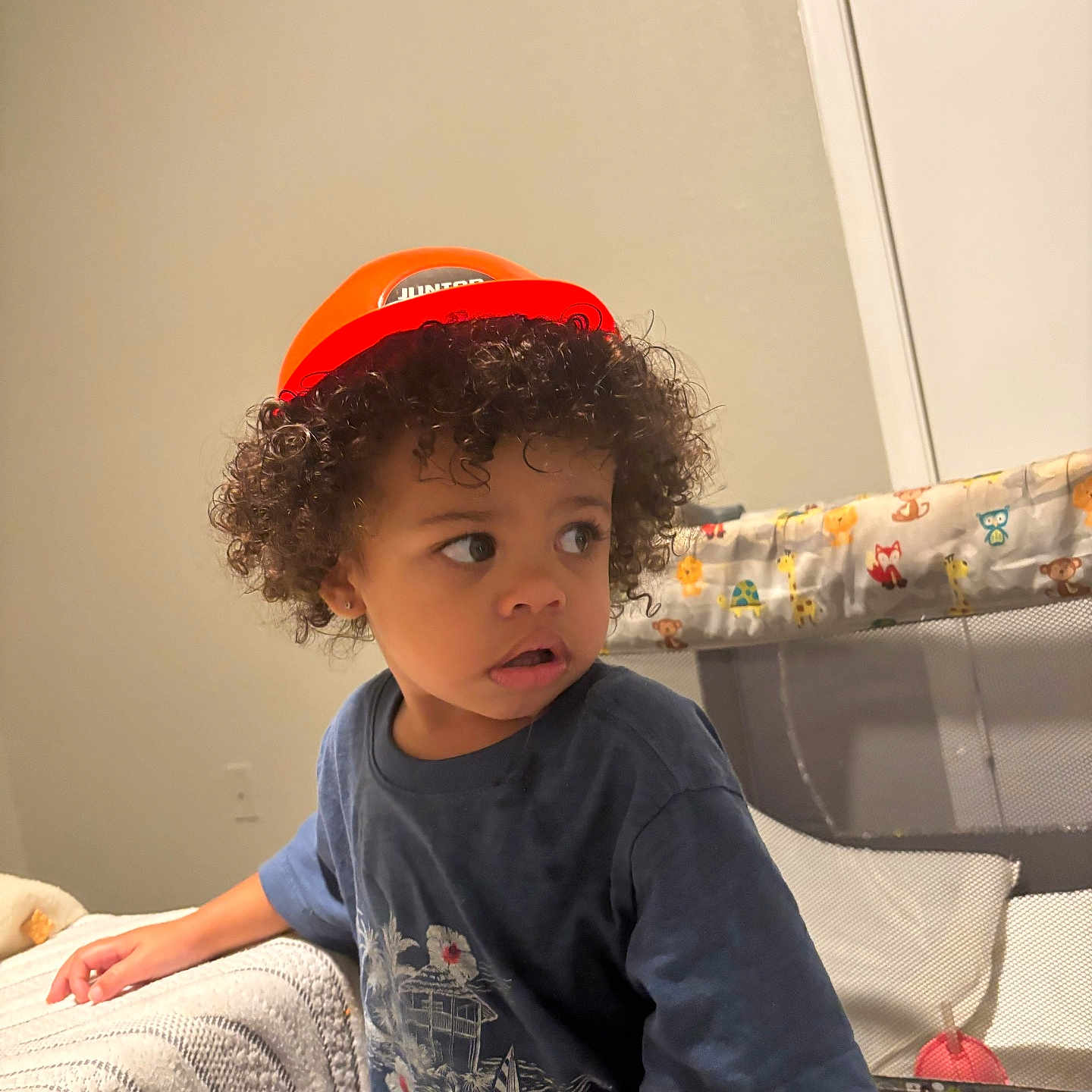 Ke-Von joined the competition — help win amazing prizes! toddler, child, curly_hair, orange_helmet, blue_shirt, indoor, playpen, blanket, animal_print, curious, looking_away, furniture, home, portrait, young_child, casual_clothing, expression, person, cute, soft_lighting