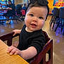 baby, child, black_clothing, headband, bow, high_chair, table, indoor, restaurant, people, background, colorful, blurred, happy, smiling, face, portrait, curious, seated, wood