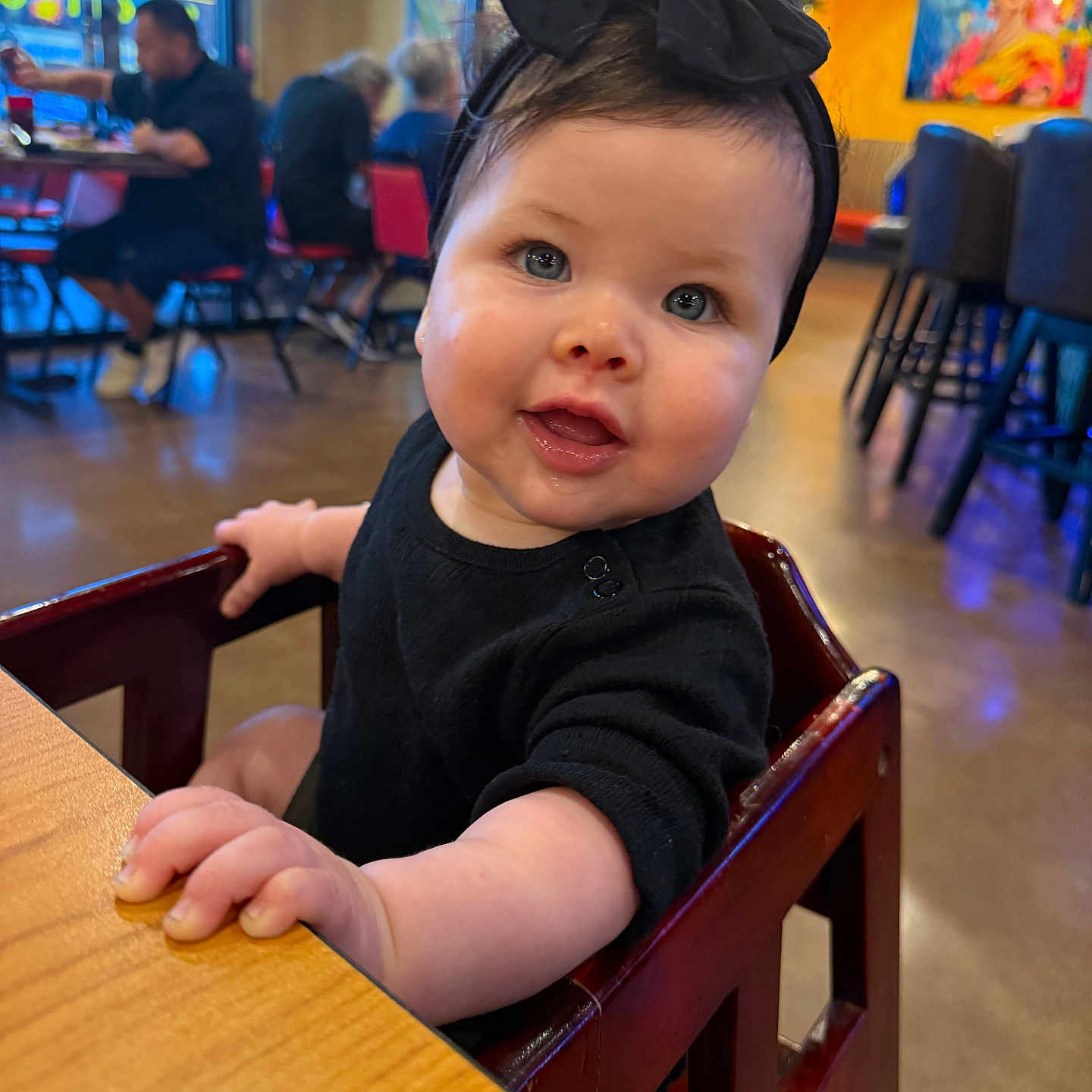 Cassandra is registered to the contest to win money with this photo: baby, background, black_clothing, blurred, bow, child, colorful, curious, face, happy, headband, high_chair, indoor, people, portrait, restaurant, seated, smiling, table, wood