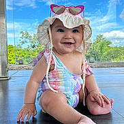 Zaenhyra is registered to the contest to win money with this photo: baby, child, smiling, sitting, floor, hat, sunglasses, heart_shaped, swimsuit, unicorn_pattern, outdoor, daylight, blue_sky, greenery, happy, cute, infant, portrait, sun_hat, barefoot