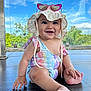 baby, child, smiling, sitting, floor, hat, sunglasses, heart_shaped, swimsuit, unicorn_pattern, outdoor, daylight, blue_sky, greenery, happy, cute, infant, portrait, sun_hat, barefoot